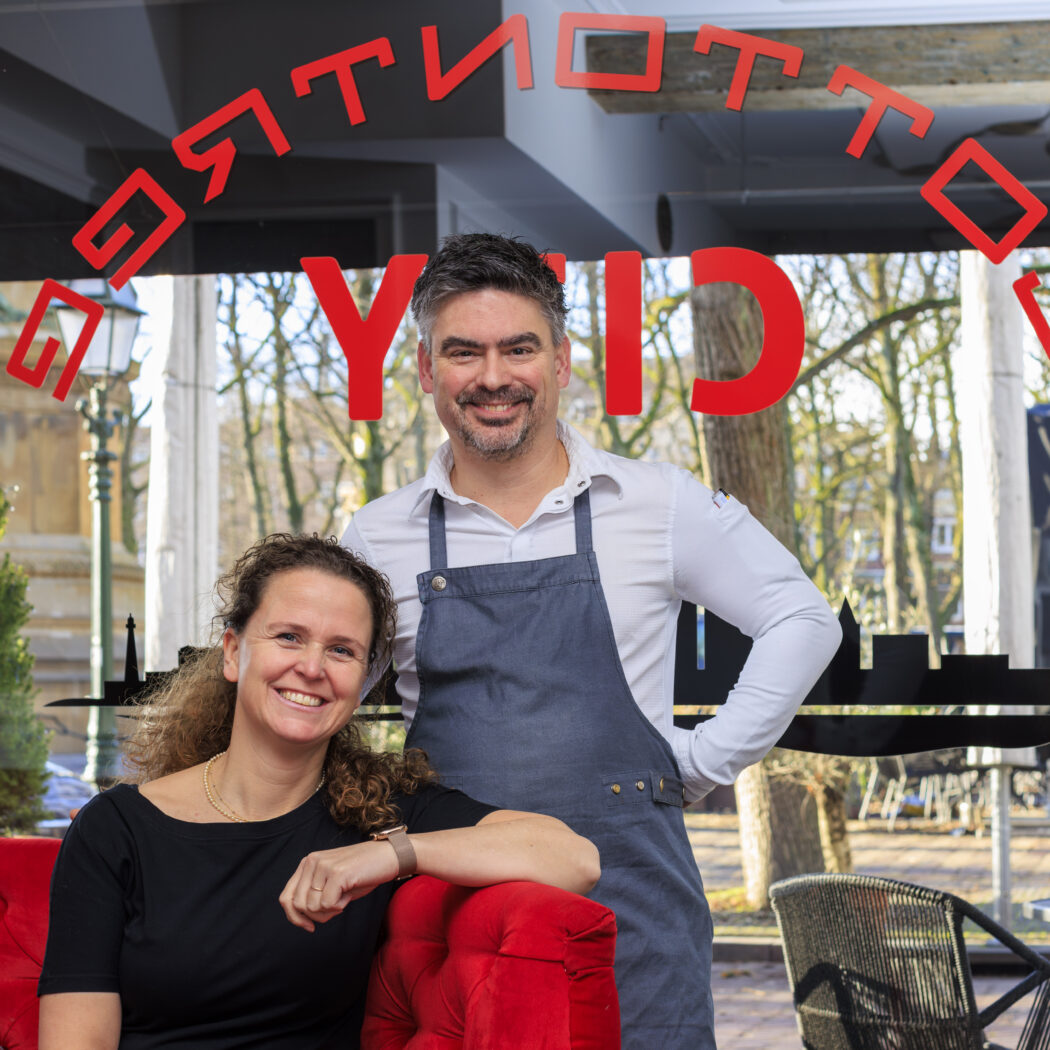 Lachende vrouw op rode bank en man met schort in restaurant voor raam met het omgekeerde TOTTI CITY.