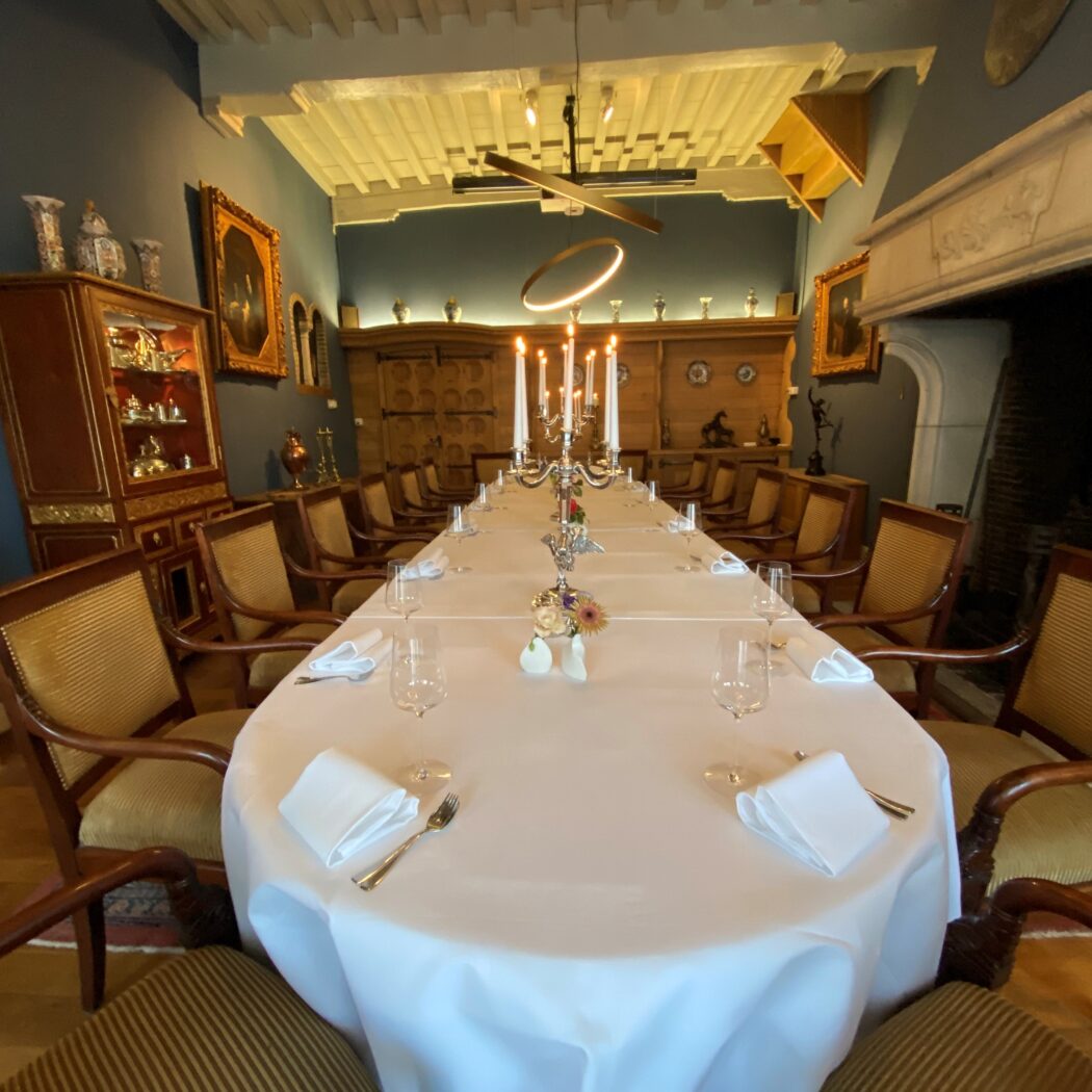 Formele eetzaal met lange gedekte tafel, zilveren kandelaars, houten stoelen en moderne verlichting aan het balkenplafond.