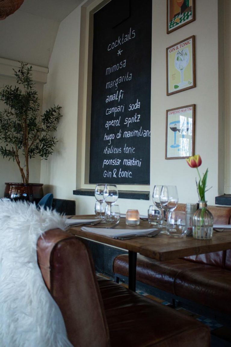 Sfeervol restaurantinterieur met gedekte houten tafel, leren banken en zwart schoolbord met cocktailmenu Mimosa en Gin & Tonic.
