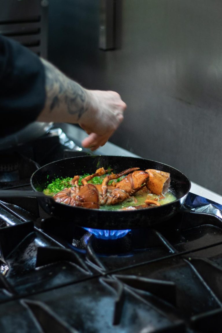 Een chef met getatoeëerde arm strooit kruiden over gekookte kreeft in een pan op een gasfornuis.