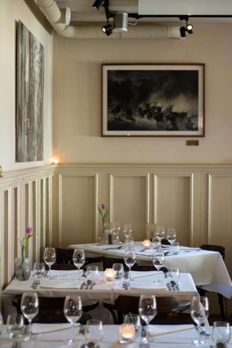 Warm verlicht restaurantinterieur met meerdere gedekte tafels, wijnglazen, kaarsen en paarse tulpen.