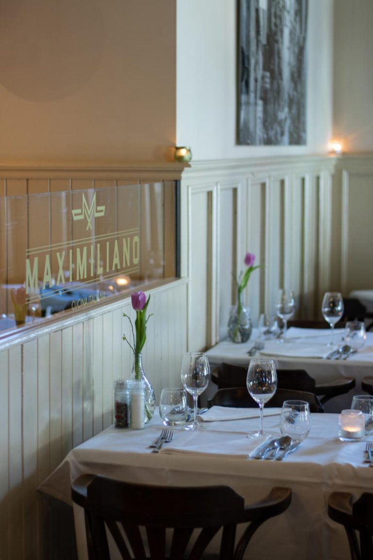 Sfeervol interieur van Italiaans restaurant Maximiliano met gedekte tafels, paarse tulpen, kaarsjes en logo.