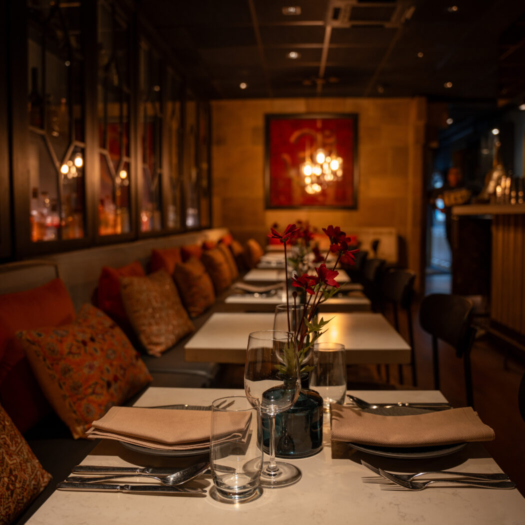 Sfeervolle restaurant eetzaal met gedekte tafels, roodbruine kussens, rode bloemen en een bar op de achtergrond.