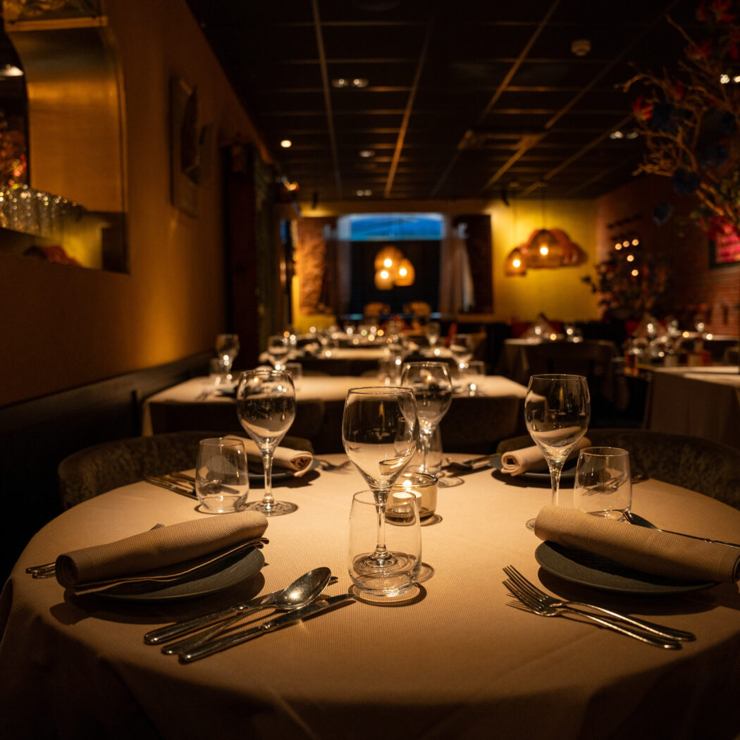 Sfeervol restaurantinterieur met meerdere elegant gedekte tafels, bestek en glazen onder warme verlichting.