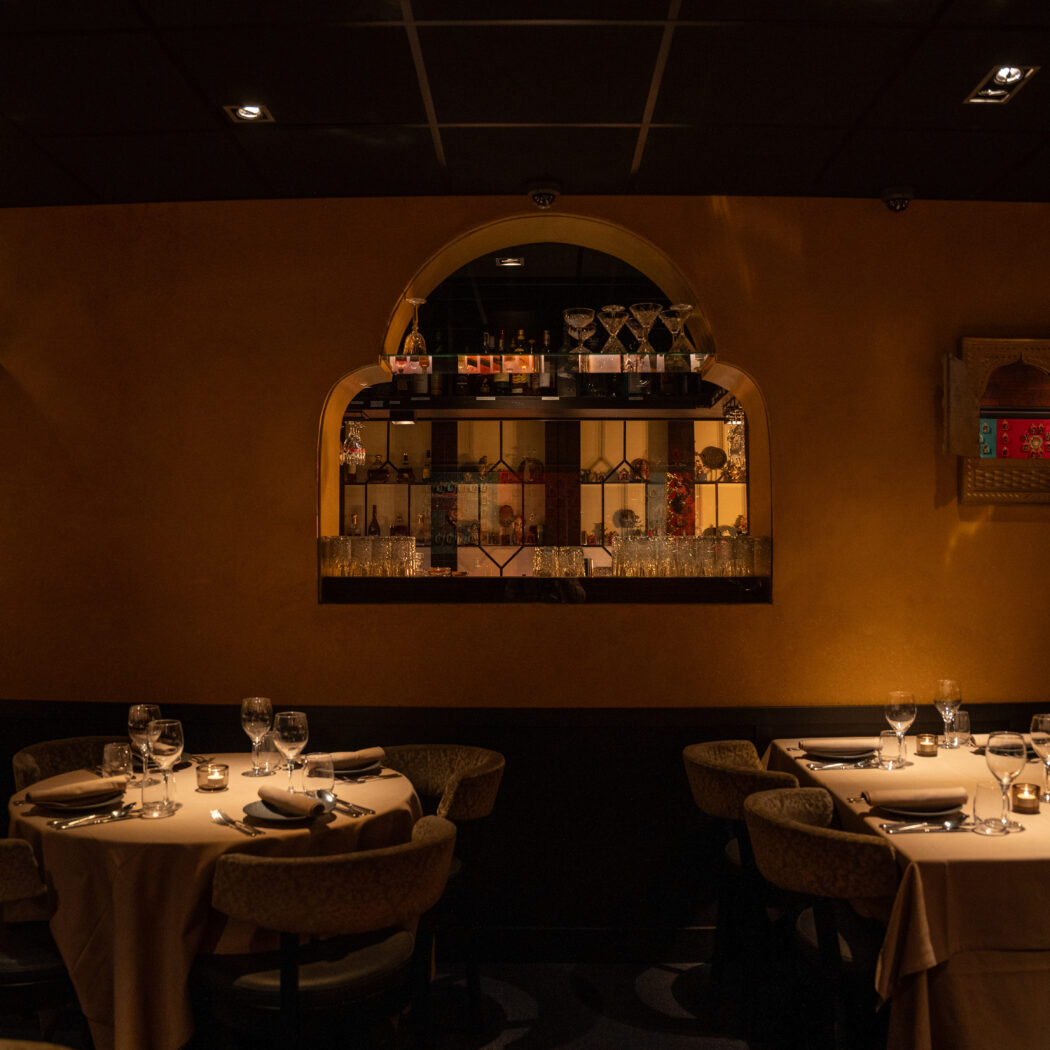 Sfeervol restaurantinterieur met warme verlichting, gedekte tafels en een nis vol flessen en glazen in een oranje muur.