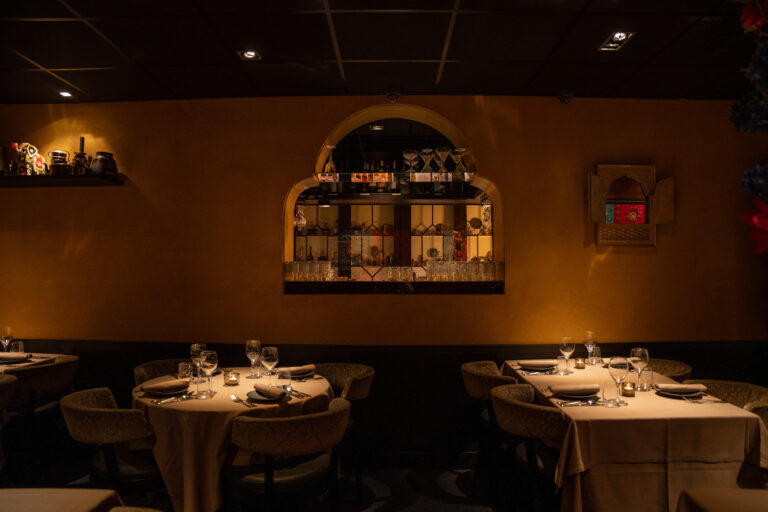 Sfeervol restaurantinterieur met warme verlichting, gedekte tafels en een nis vol flessen en glazen in een oranje muur.