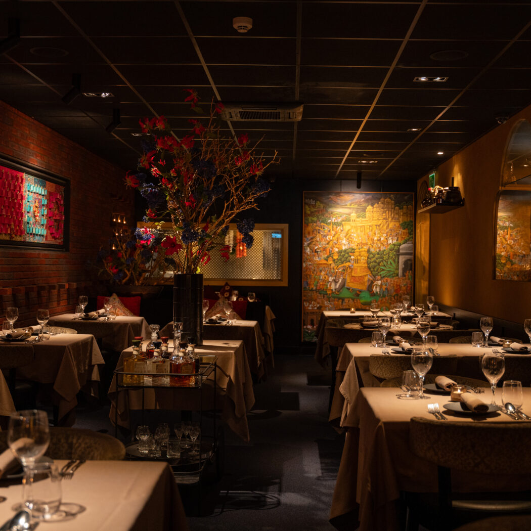 Sfeervol restaurantinterieur met gedekte tafels, bakstenen muur, oosterse kunst en grote bloemdecoratie.