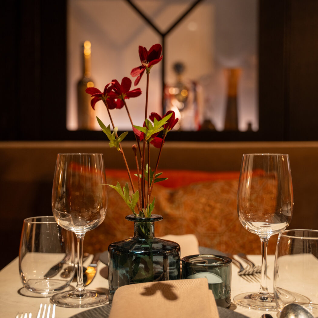 Sfeervol gedekte restauranttafel voor twee met donkerrode bloemen, kaarslicht en wijnglazen.