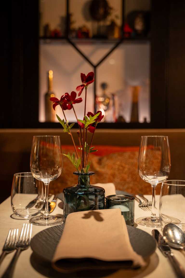 Sfeervol gedekte restauranttafel voor twee met donkerrode bloemen, kaarslicht en wijnglazen.
