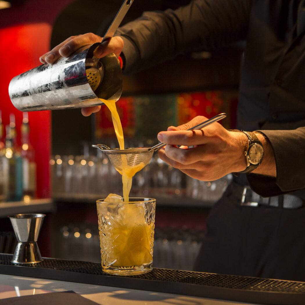 Barman schenkt geel drankje uit cocktailshaker door zeef in een gedetailleerd glas met ijs.