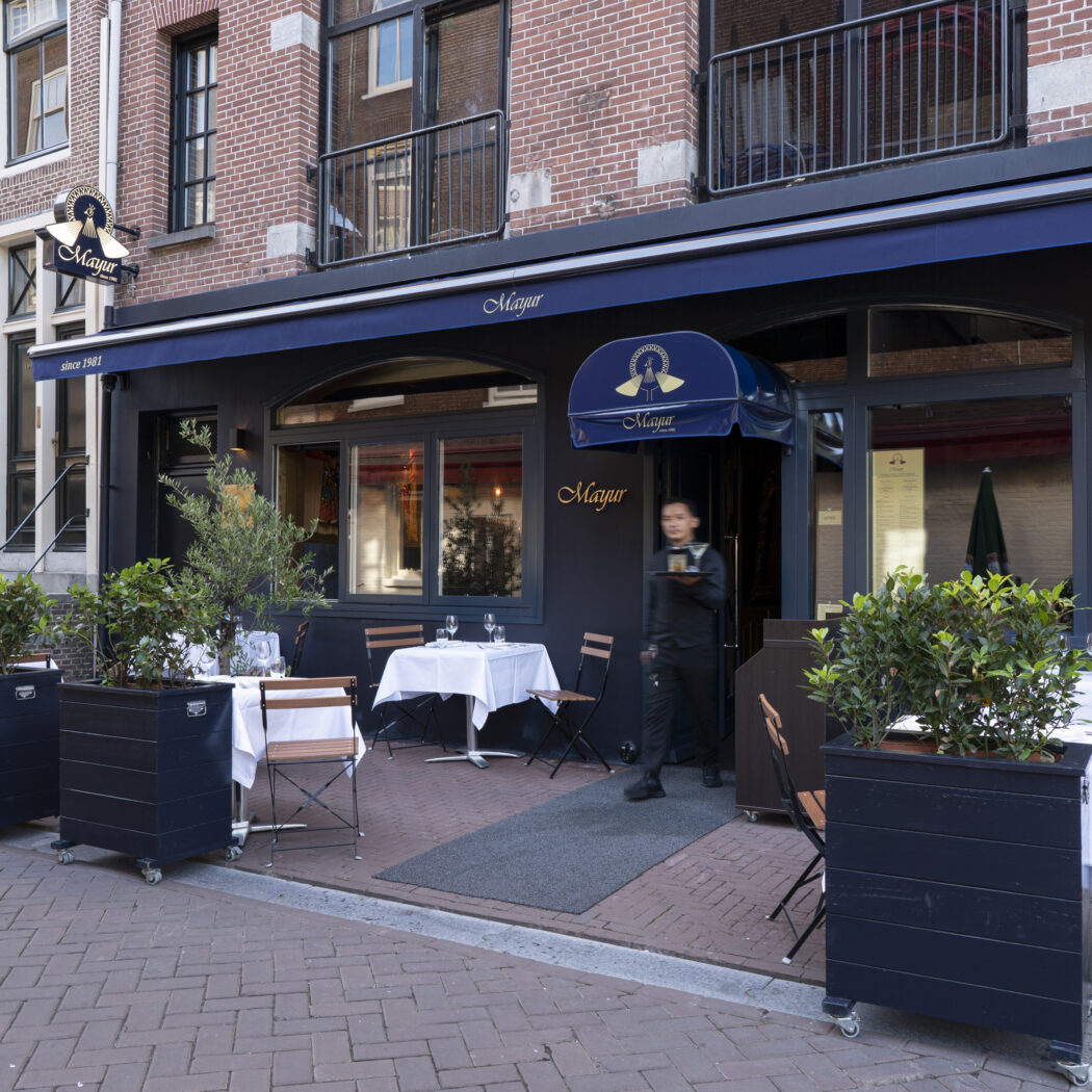 Indiaas fine dining restaurant Mayur met blauwe luifels en terras, een waiter komt naar buiten met een dienblad.