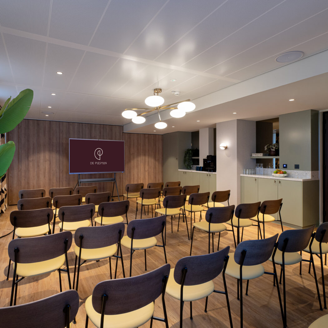 Moderne vergaderruimte met rijen houten stoelen met gele zittingen, scherm met DE PUESMAN en verfrissingshoek.