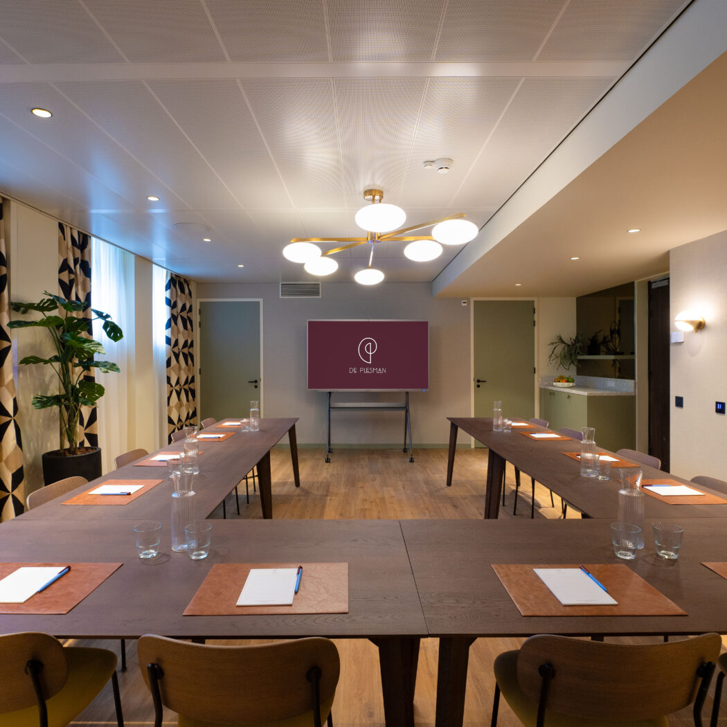 Moderne vergaderzaal met U-vormige tafel, presentatiescherm met “DE PIESMAN” logo, en een koffiebar.
