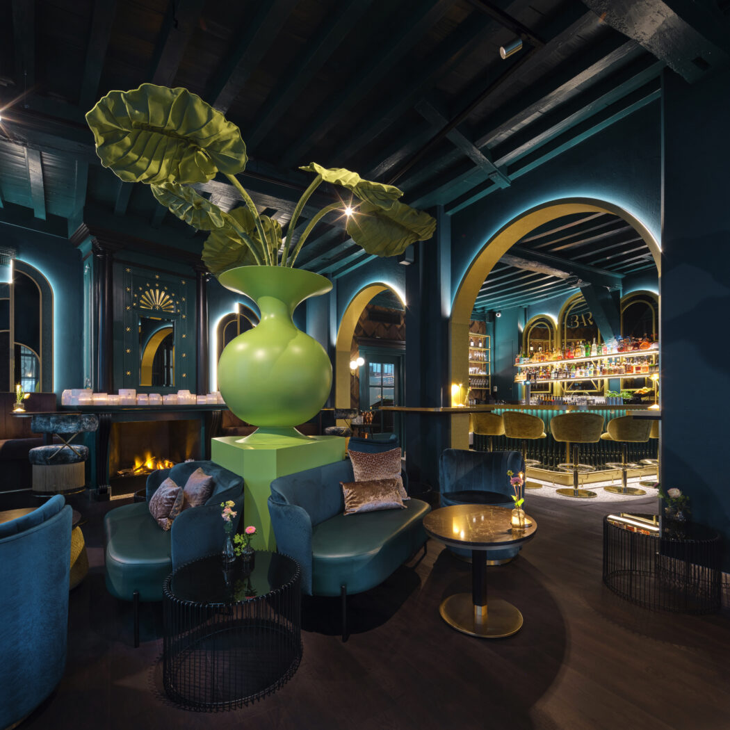 Stijlvolle bar en lounge met grote groene vaas, donkergroene fluwelen zitjes, open haard en verlichte bar met BR-logo.