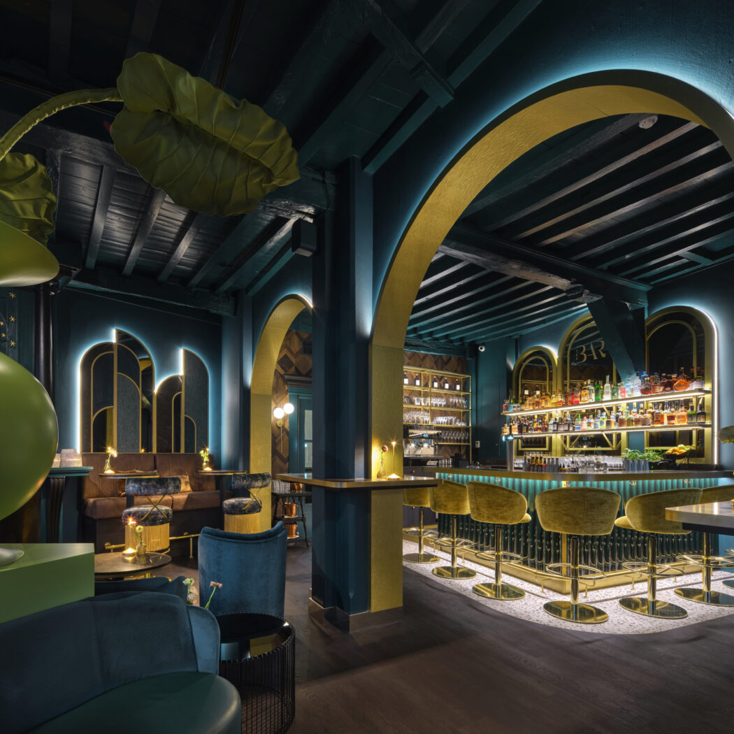 Luxe loungebar interieur met blauwgroene muren, gouden bogen, verlichte bar, gele krukken en grote groene decoratieve vaas.