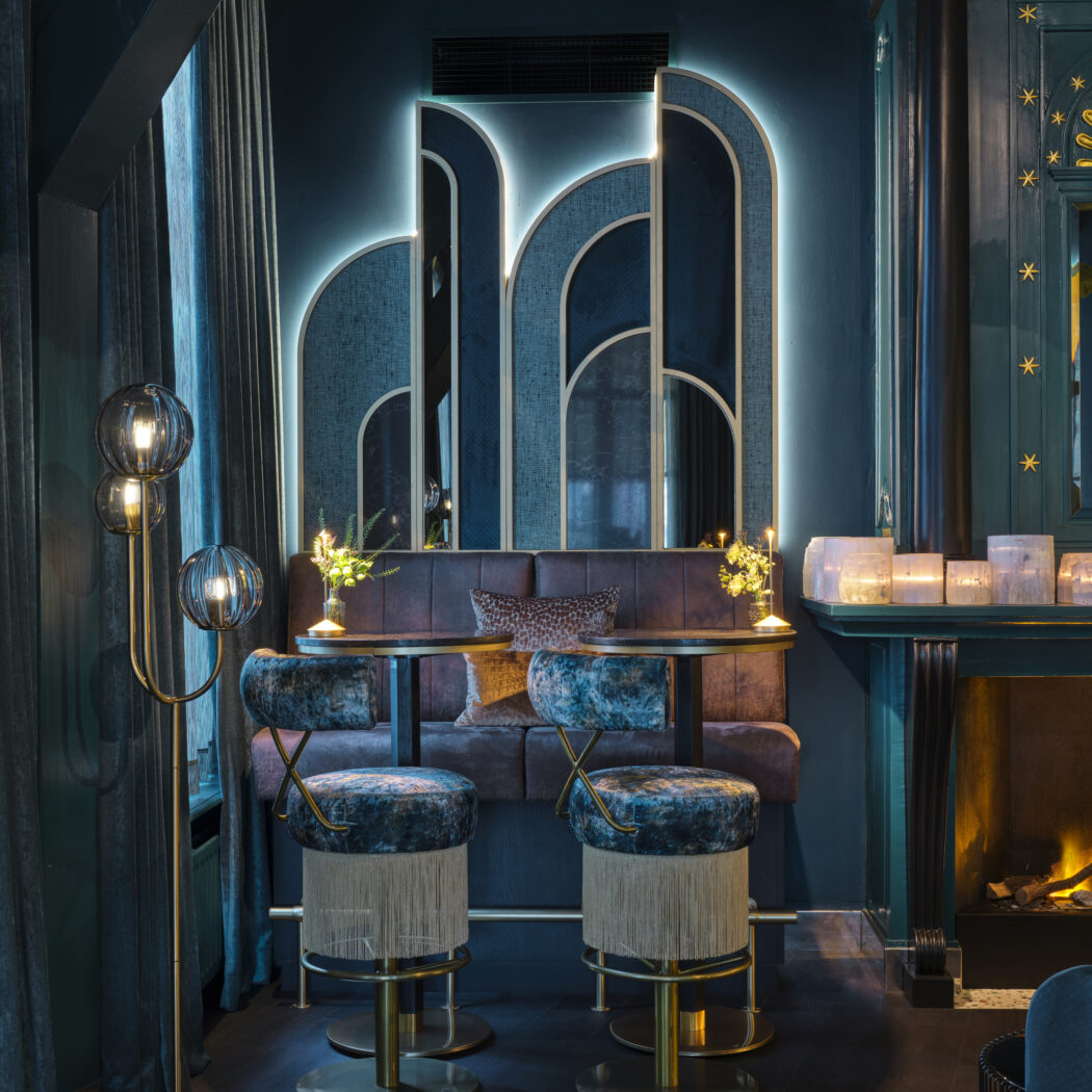 Luxe donkerblauwe lounge met fluwelen barkrukken, art deco spiegelwand en sfeervolle verlichting.