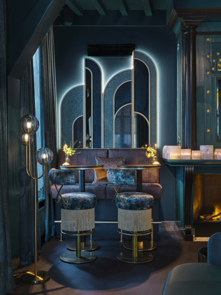 Luxe donkerblauwe lounge met fluwelen barkrukken, art deco spiegelwand en sfeervolle verlichting.