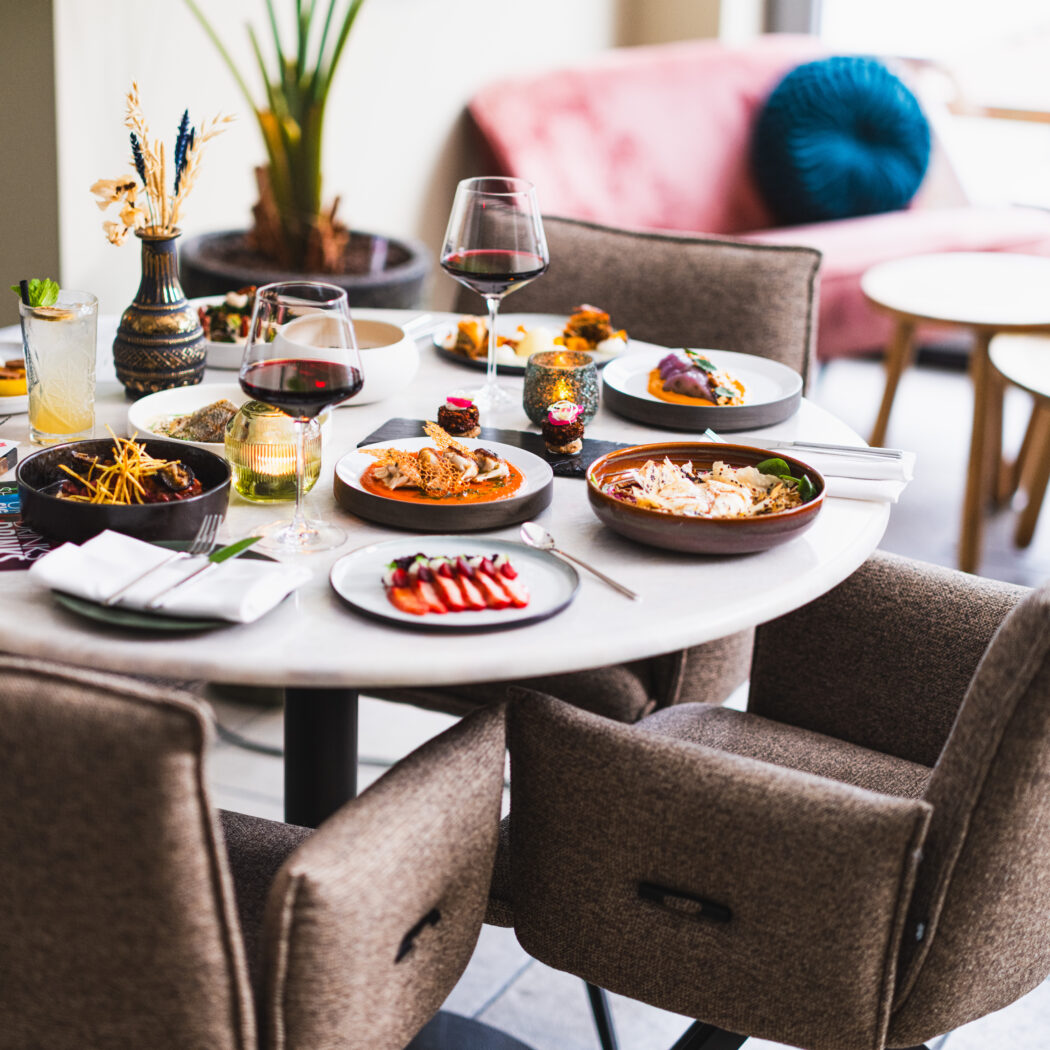 Ronde witte tafel met diverse kleine gerechten, wijnglazen en een cocktail in een sfeervol restaurant.
