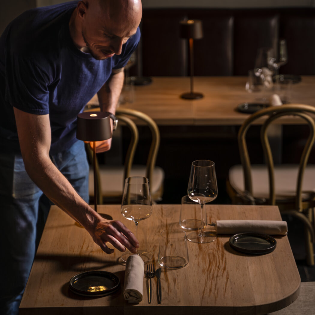 Man dekt een houten restauranttafel met wijnglazen en bestek in een sfeervol verlichte ruimte.