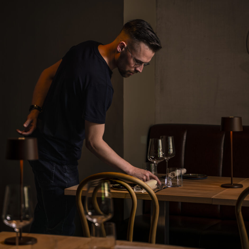 Man in donker T-shirt schikt servet en bestek op houten tafel in een sfeervol restaurant.