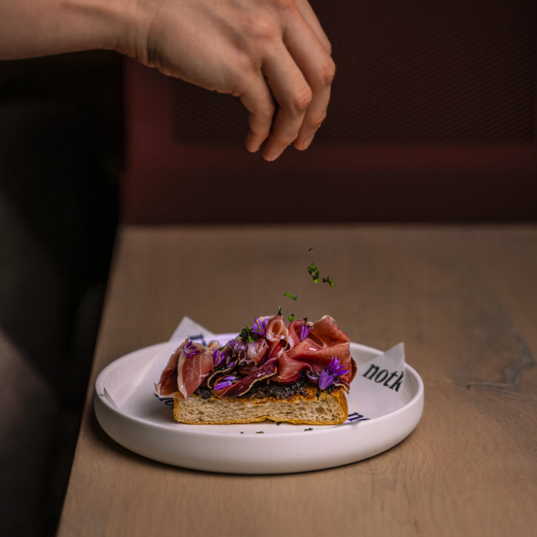 Een hand strooit verse kruiden over een gourmet broodje met prosciutto, tapenade en bieslookbloemen op een wit bord.
