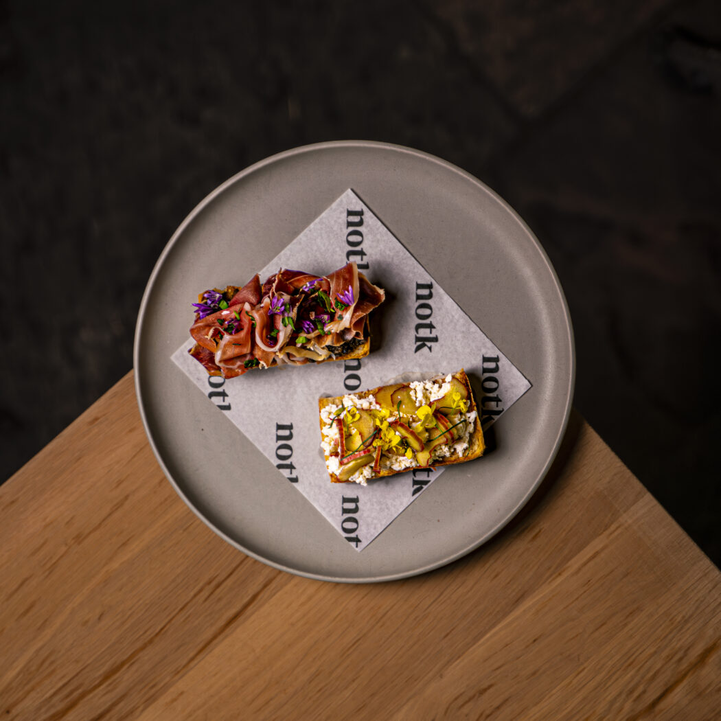 Luxe belegde broodjes met prosciutto, roomkaas, fruit en eetbare bloemen op grijs bord met notk-papier.