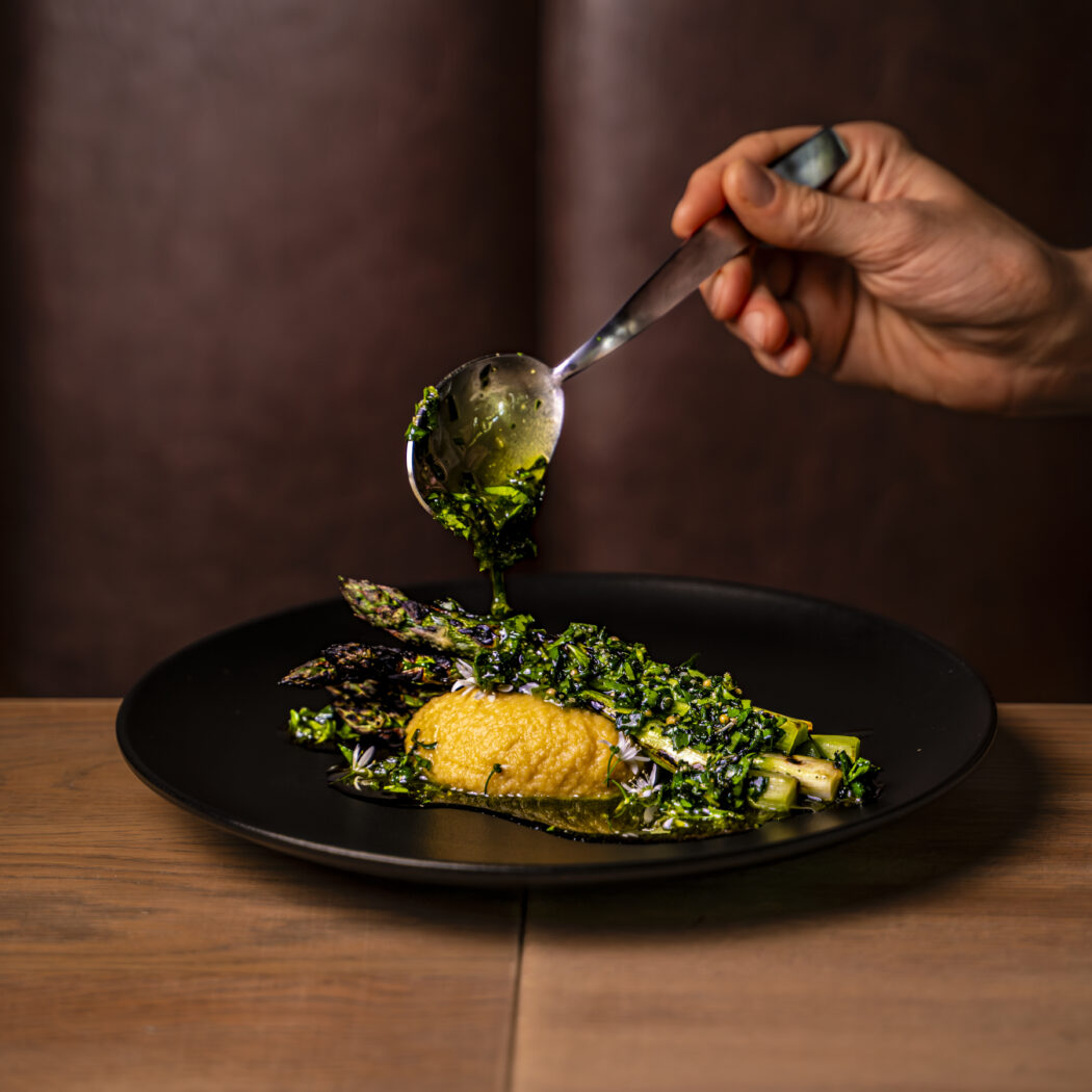 Een hand giet groene kruidenolie over gegrilde asperges, gele puree en verse kruiden op een zwarte schaal.