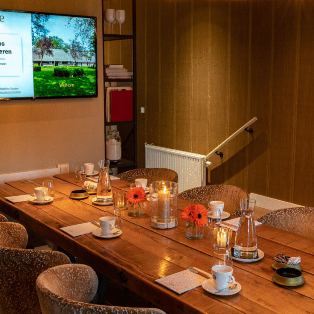 Lange houten vergadertafel gedekt met koffie, water, oranje bloemen en kaarsen, met tv-scherm Draadloos presenteren.