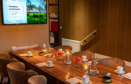 Lange houten vergadertafel gedekt met koffie, water, oranje bloemen en kaarsen, met tv-scherm Draadloos presenteren.