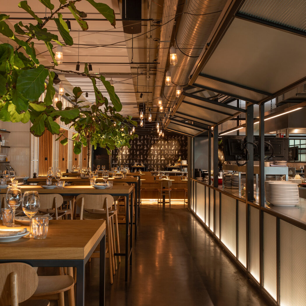Warm verlicht modern restaurantinterieur met gedekte houten tafels, groene planten en open keuken.