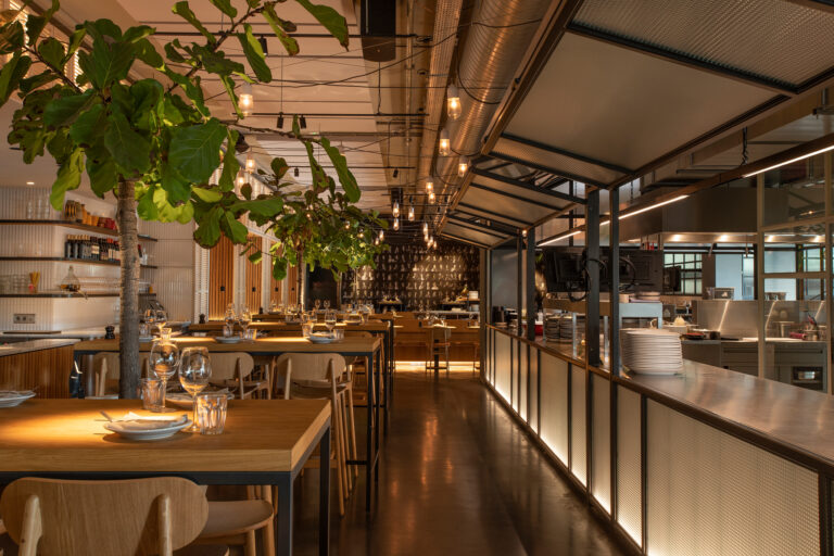 Warm verlicht modern restaurantinterieur met gedekte houten tafels, groene planten en open keuken.