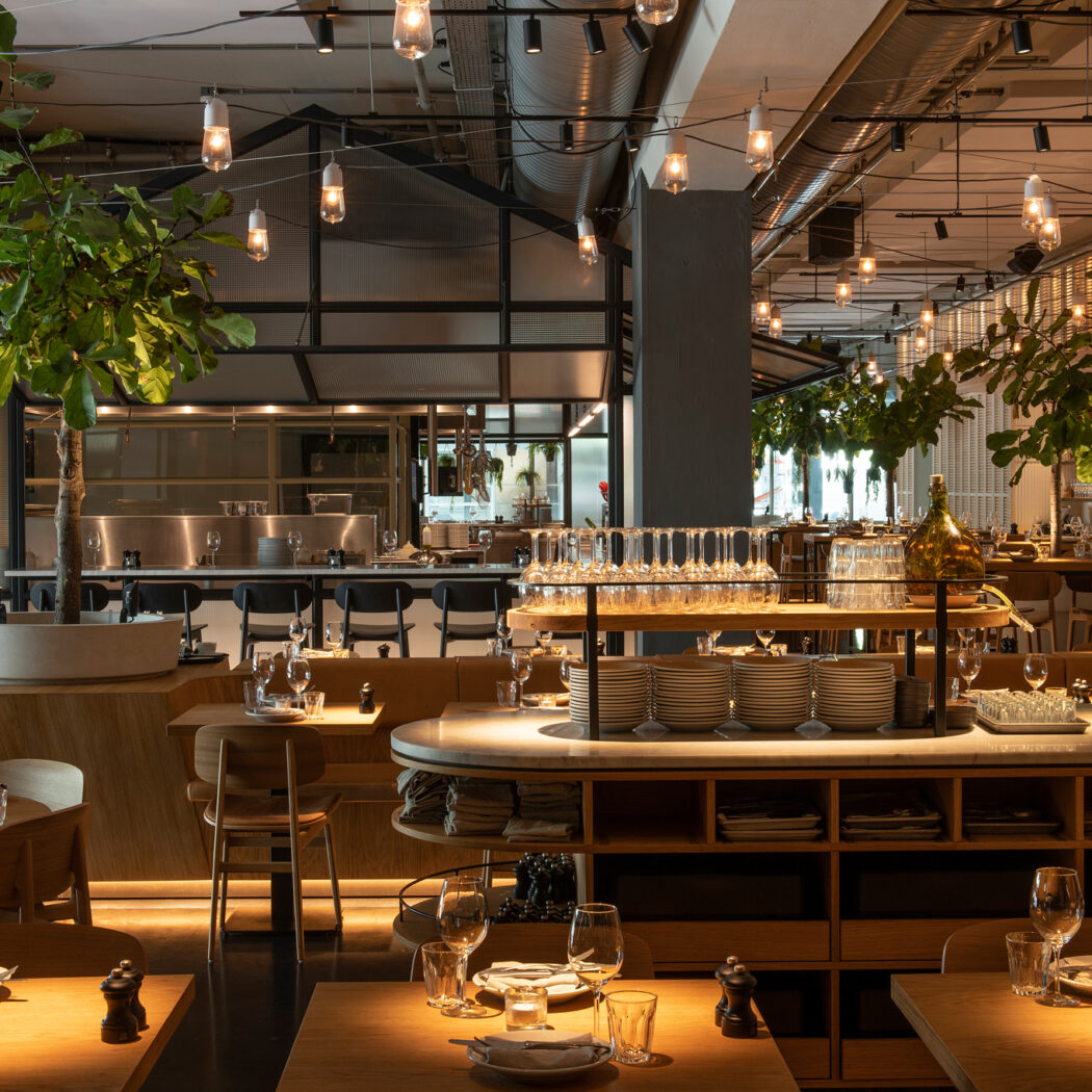 Modern restaurant interieur met opgedekte houten tafels, sfeervolle hanglampen en groene planten.