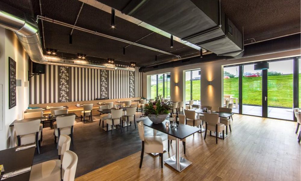Modern restaurant interior with black ceiling, striped walls, beige chairs and tables, and large windows overlooking greenery.