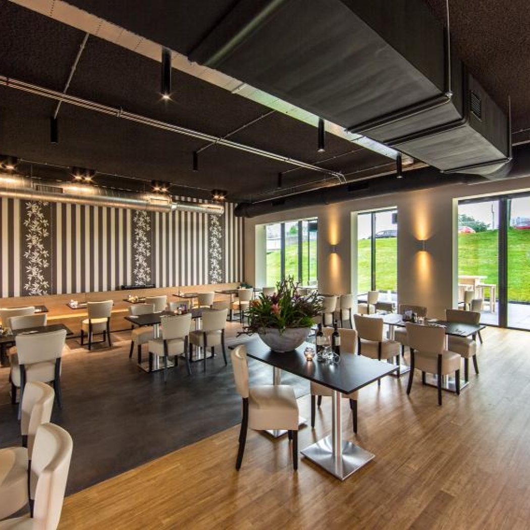 Modern restaurant interieur met zwart plafond, gestreepte muren, beige stoelen en tafels, en grote ramen met uitzicht op groen.