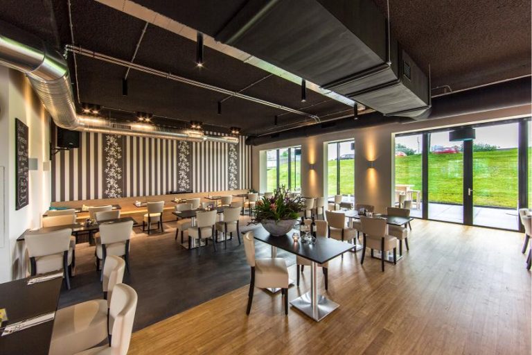 Modern restaurant interieur met zwart plafond, gestreepte muren, beige stoelen en tafels, en grote ramen met uitzicht op groen.