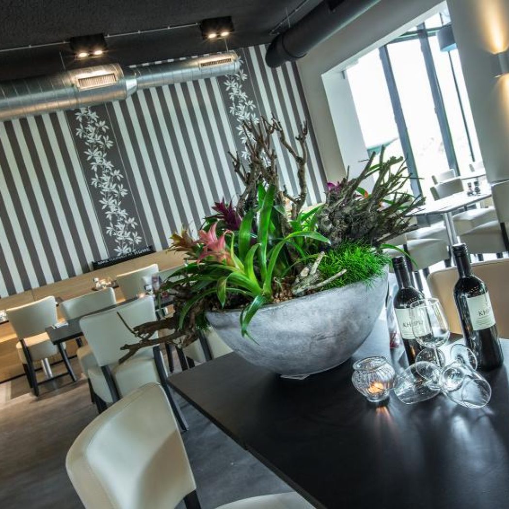 Sfeervol restaurantinterieur met grote decoratieve plantenbak, wijnflessen en glazen op donkere tafel.