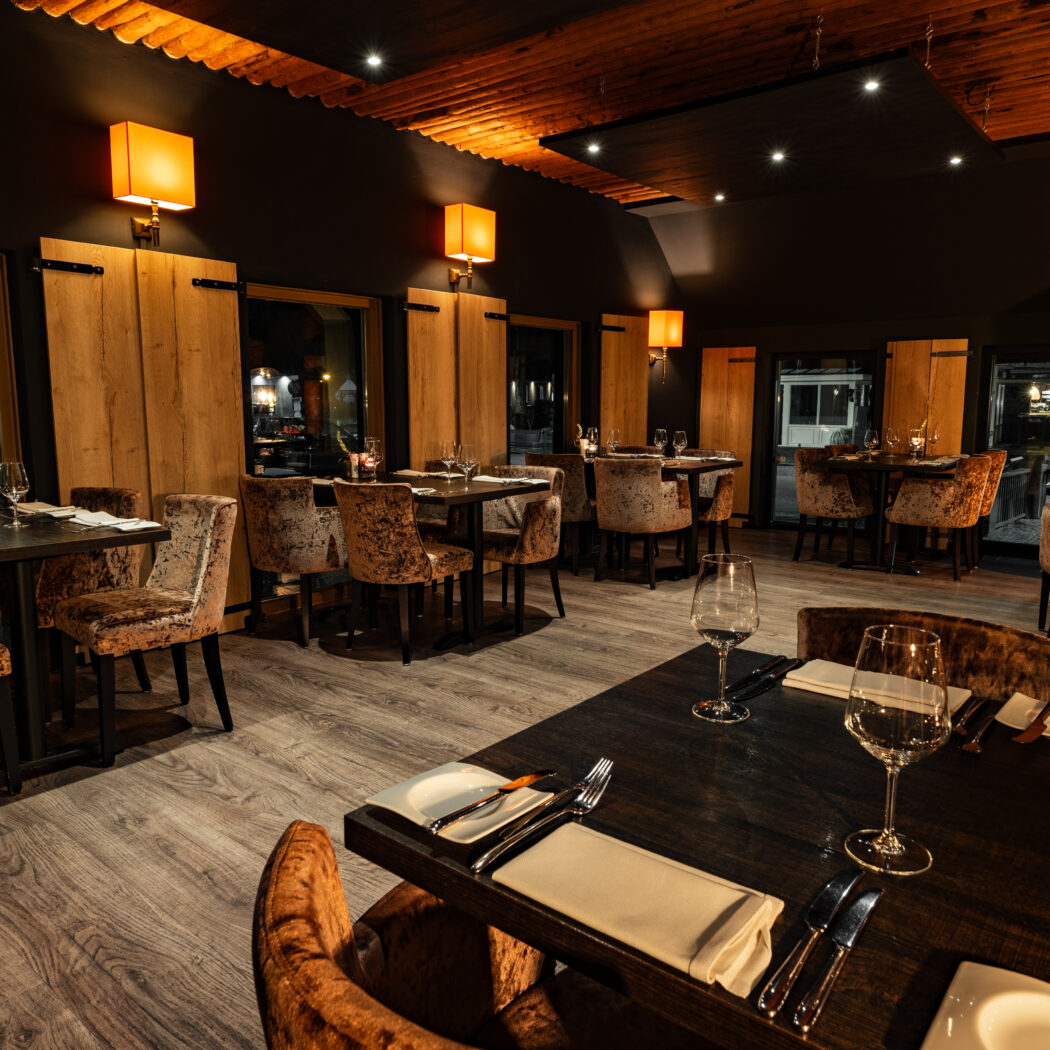 Sfeervol restaurant interieur met gedekte tafels, bruine fluwelen stoelen, houten accenten en warme verlichting.