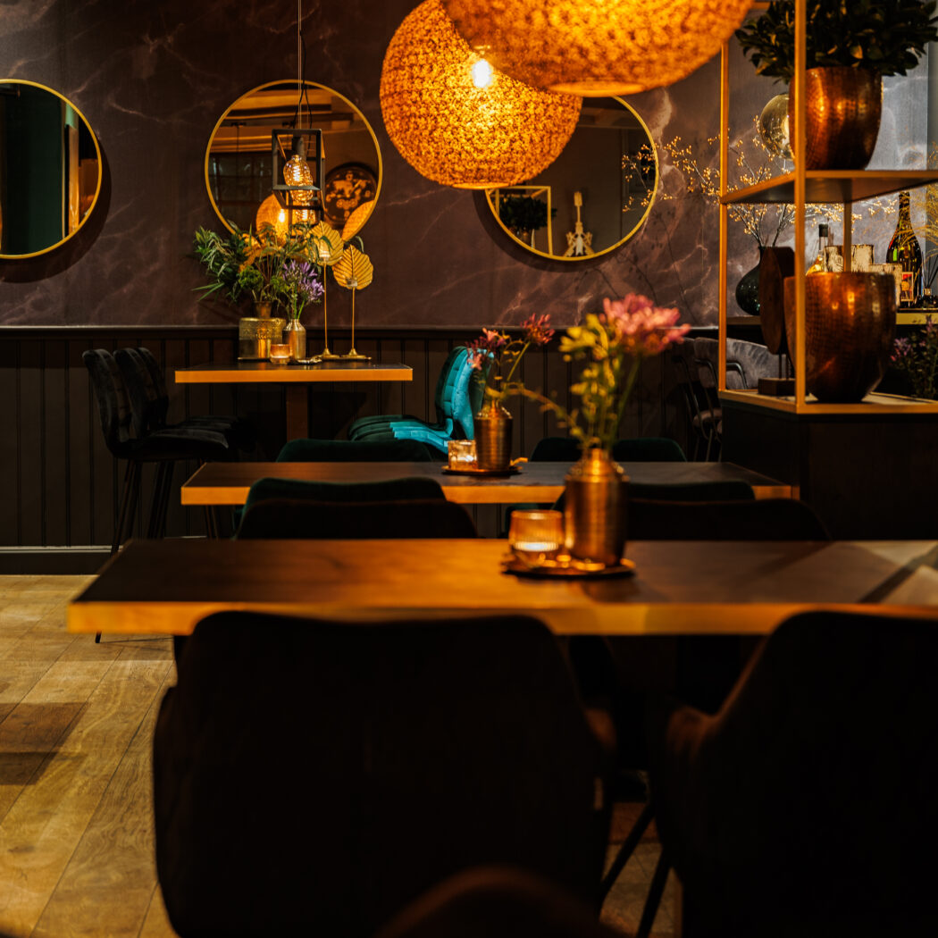 Sfeervol restaurantinterieur bij avond met oranje hanglampen, houten tafels en ronde gouden spiegels.