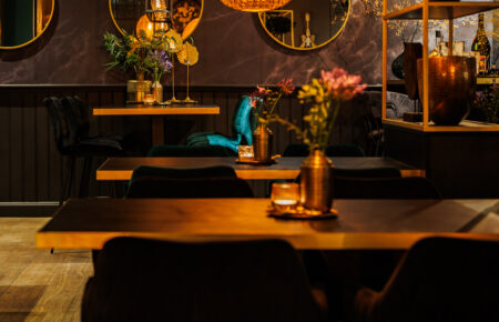 Sfeervol restaurantinterieur bij avond met oranje hanglampen, houten tafels en ronde gouden spiegels.