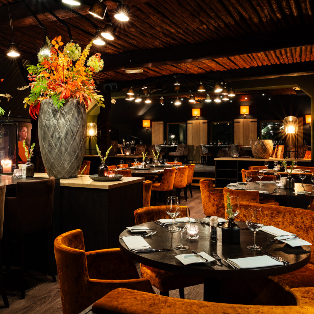 Sfeervol restaurant met houten plafond, oranje fluwelen zitplaatsen en grote bloemstukken.
