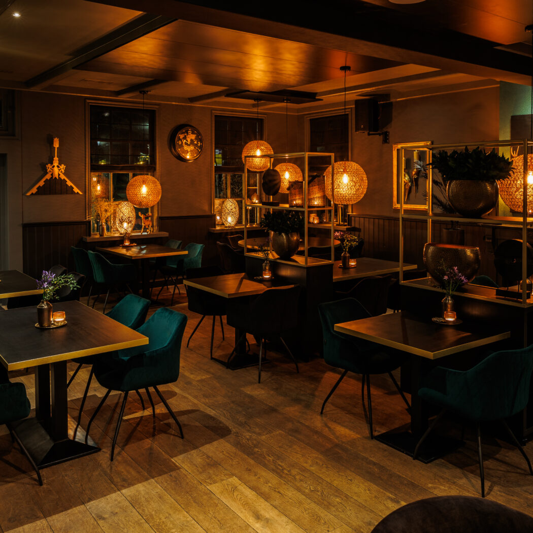Sfeervol restaurant met donkere tafels, smaragdgroene stoelen, houten vloer en een glazen wijnkoeler.