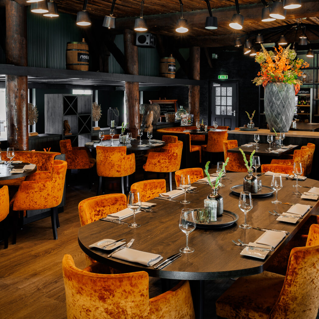 Sfeervol restaurantinterieur met oranje fluwelen stoelen, gedekte houten tafels, bloemen en wijnvaten.