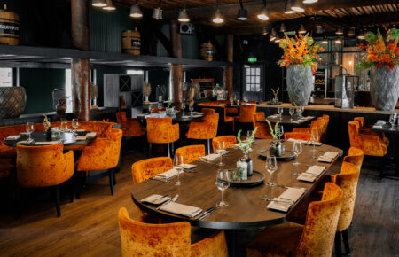 Sfeervol restaurantinterieur met oranje fluwelen stoelen, gedekte houten tafels, bloemen en wijnvaten.