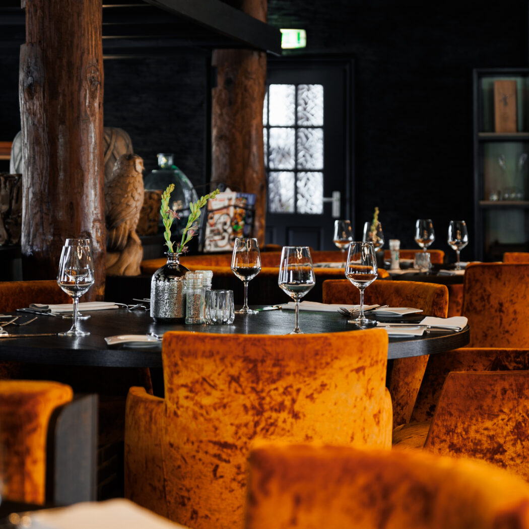 Sfeervol restaurant met gedekte tafels, oranje fluwelen stoelen en robuuste houten pilaren.