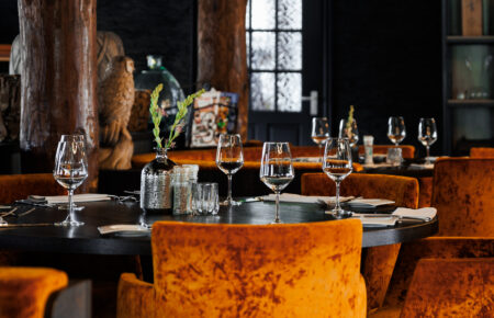 Sfeervol restaurant met gedekte tafels, oranje fluwelen stoelen en robuuste houten pilaren.