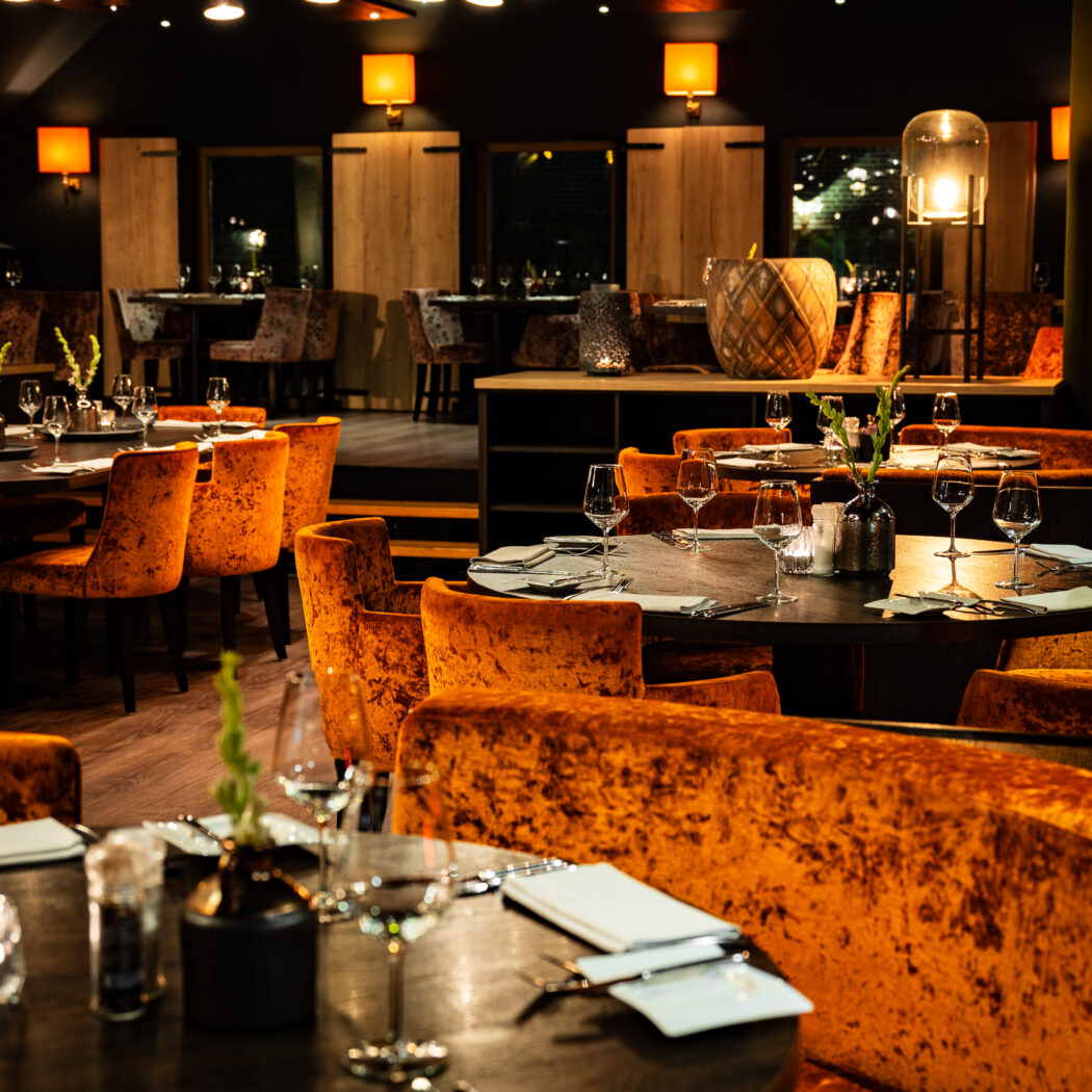 Sfeervol restaurantinterieur met ronde tafels, oranje fluwelen stoelen en banken, verlicht door warm sfeerlicht.