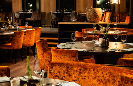 Sfeervol restaurantinterieur met ronde tafels, oranje fluwelen stoelen en banken, verlicht door warm sfeerlicht.