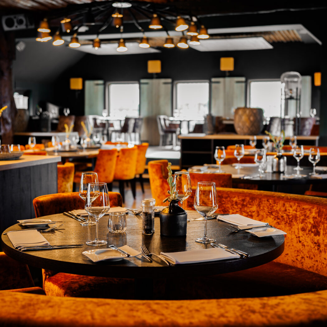Sfeervol restaurant met ronde zwarte tafels, oranje fluwelen zitbanken en gedekte plaatsen in warme tinten.