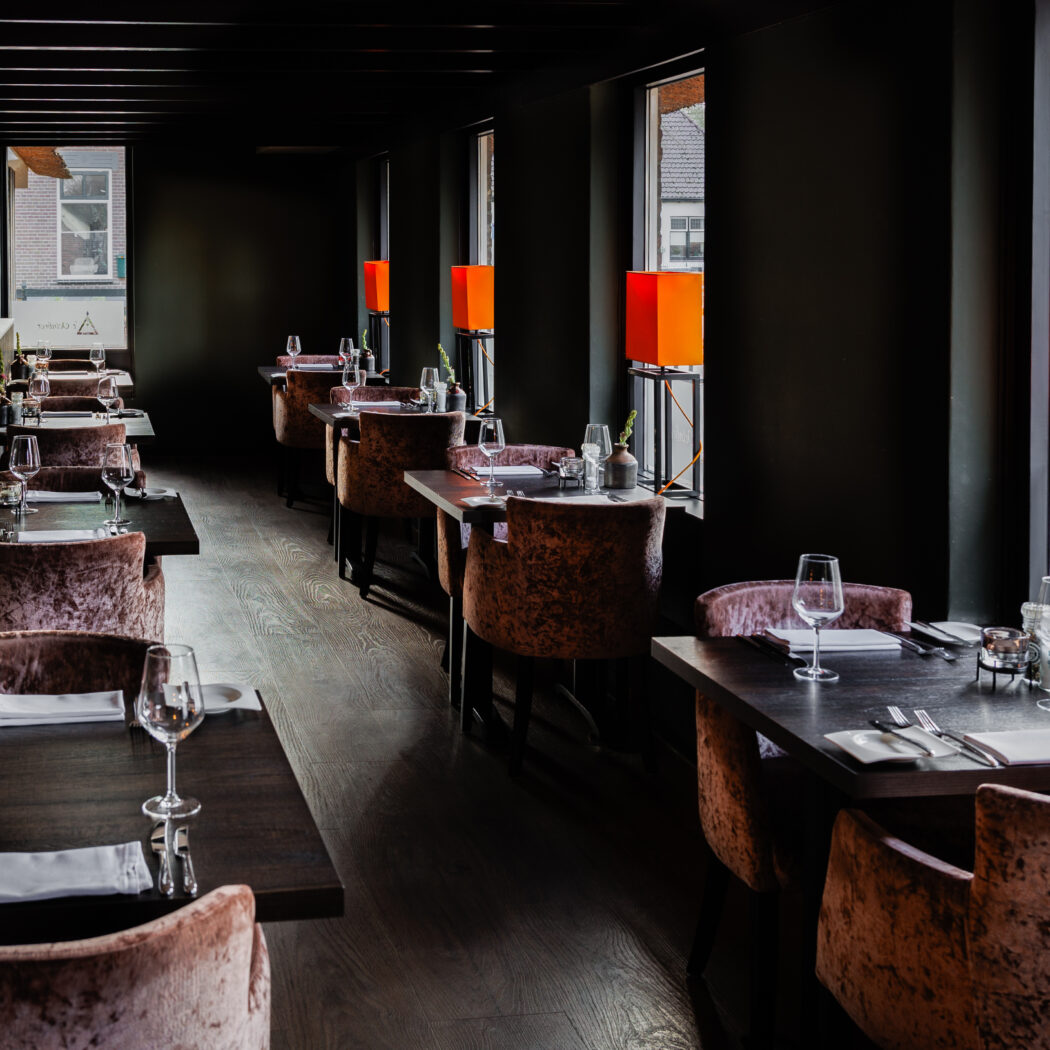 Sfeervol restaurant met donkere houten vloeren, roze fluwelen stoelen en gedekte tafels bij ramen met oranje lampen.