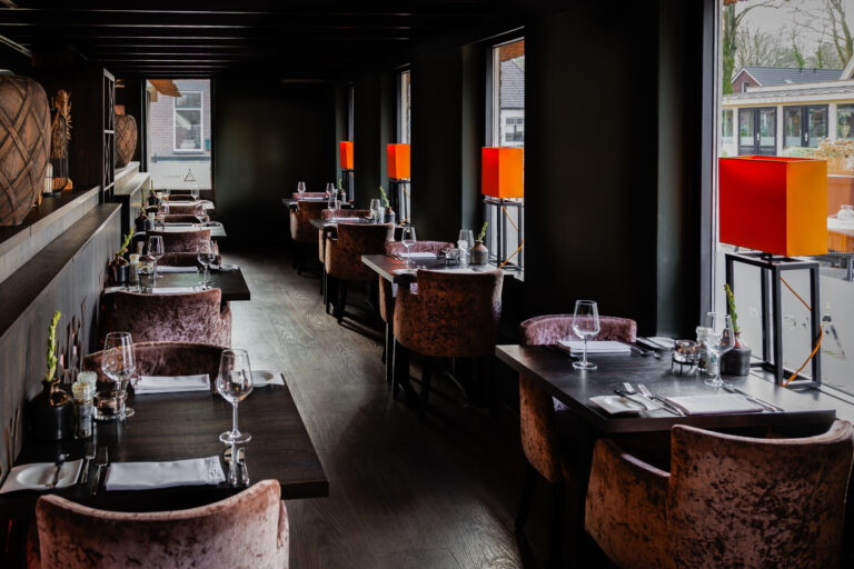 Sfeervol restaurant met donkere houten vloeren, roze fluwelen stoelen en gedekte tafels bij ramen met oranje lampen.
